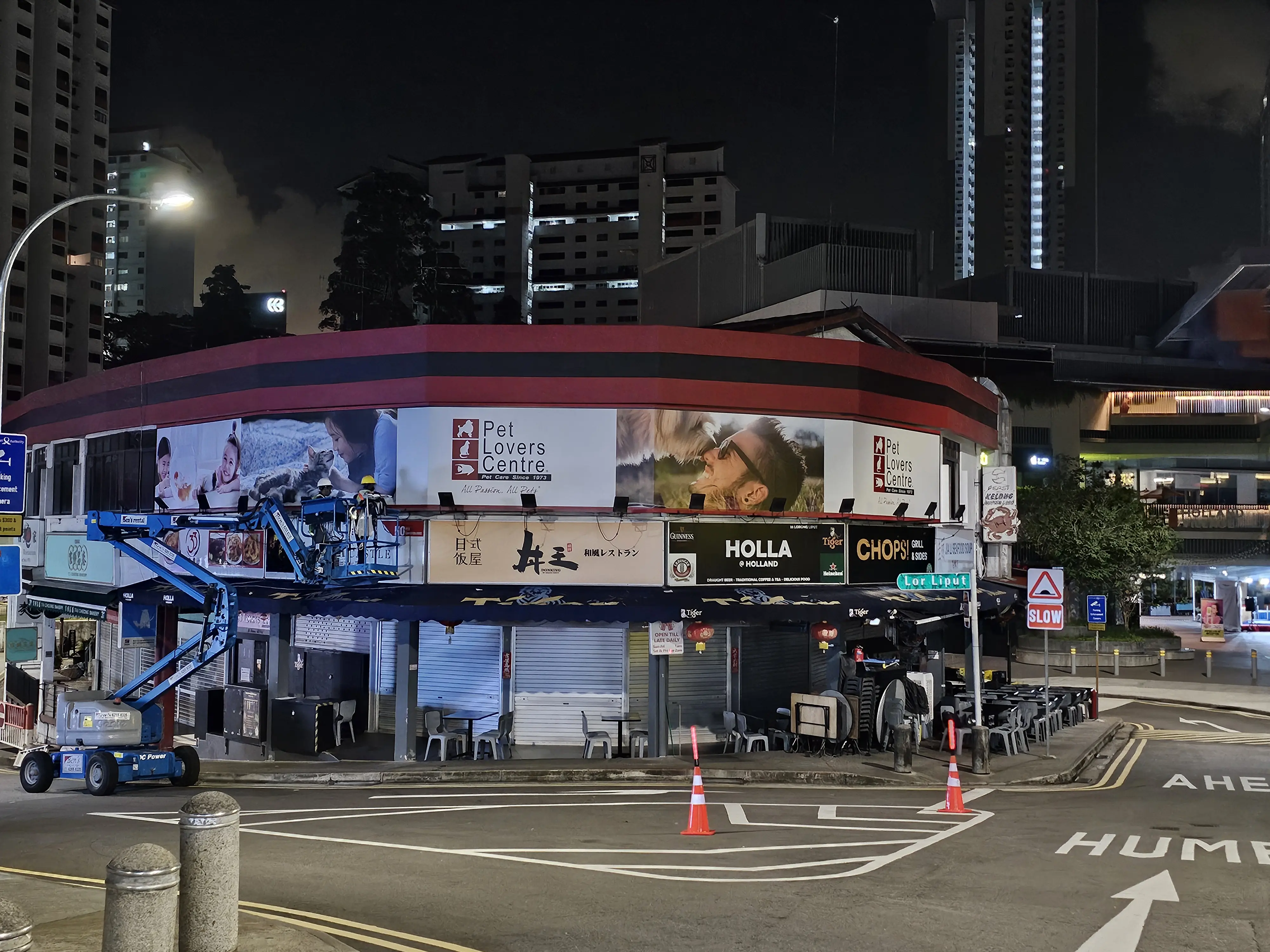 Night view of a commercial building in Singapore with shops like Pet Lovers Centre, Holla @ Holland, and CHOPS! Grill & Sides; workers on a boom lift updating signage under well-lit surroundings, showcasing the durability and importance of waterproof sticker printing for outdoor branding.