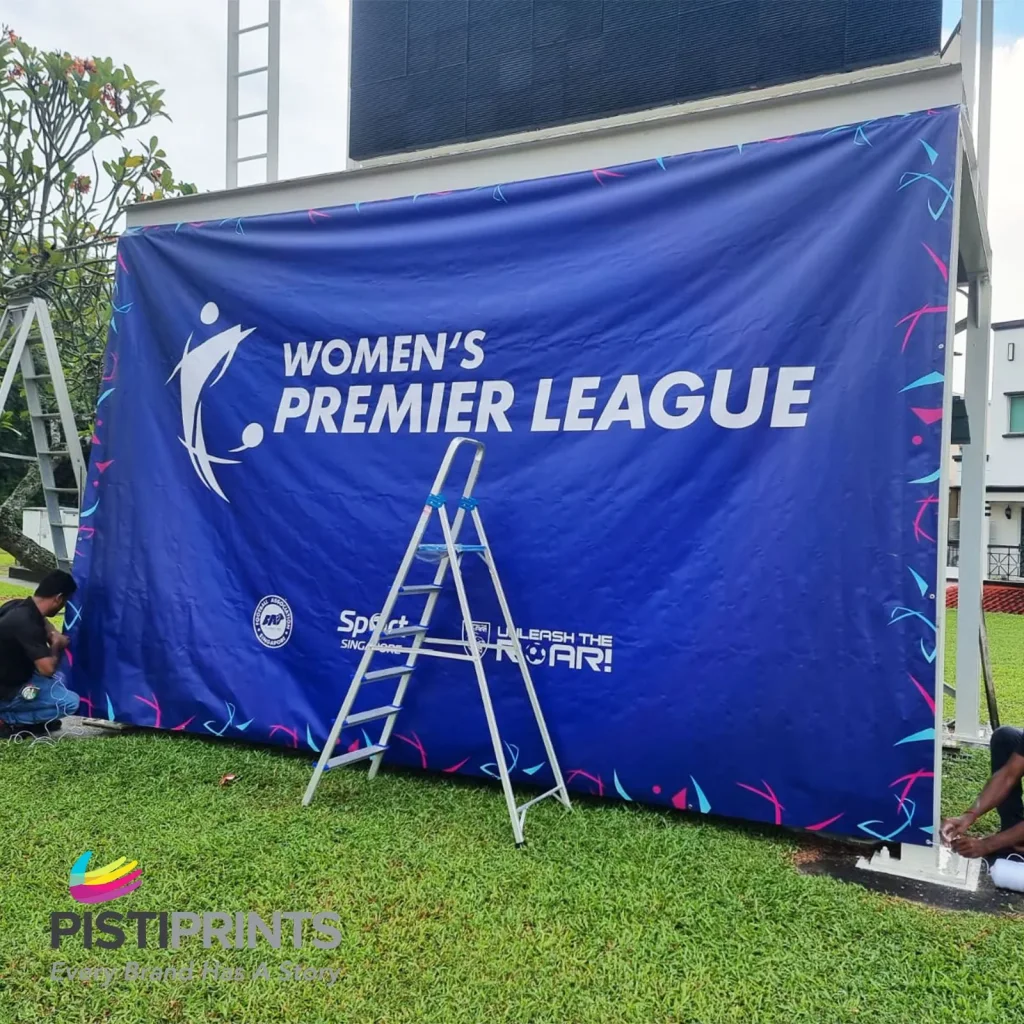Technicians setting up a bold blue 'Women's Premier League' banner outdoors, showcasing eye-catching design and professional banner printing by Pistiprints in Singapore.