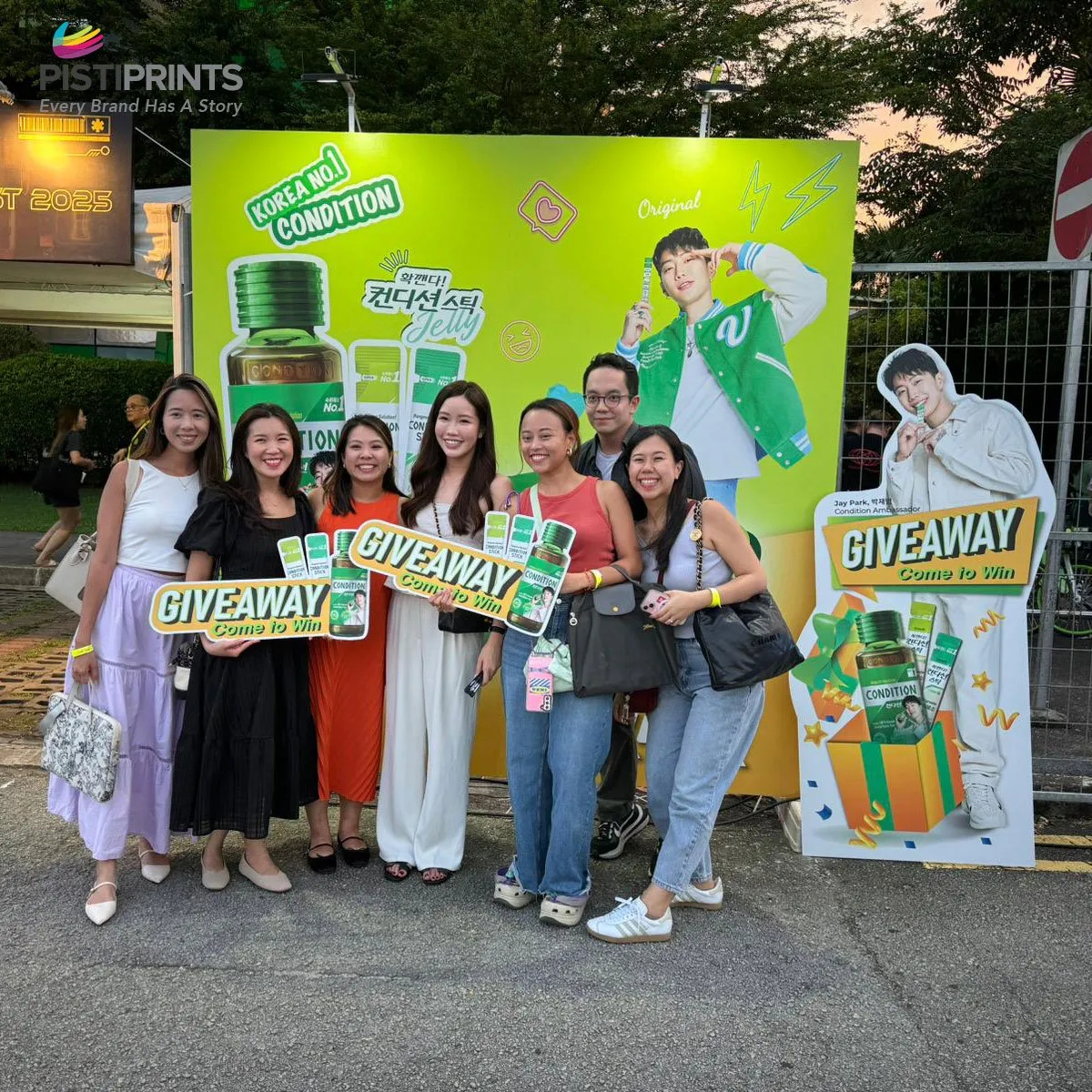 Group of people posing in front of a bright green promotional booth for Korean Condition drink, holding giveaway signs at an outdoor event organized by Pisti Prints.