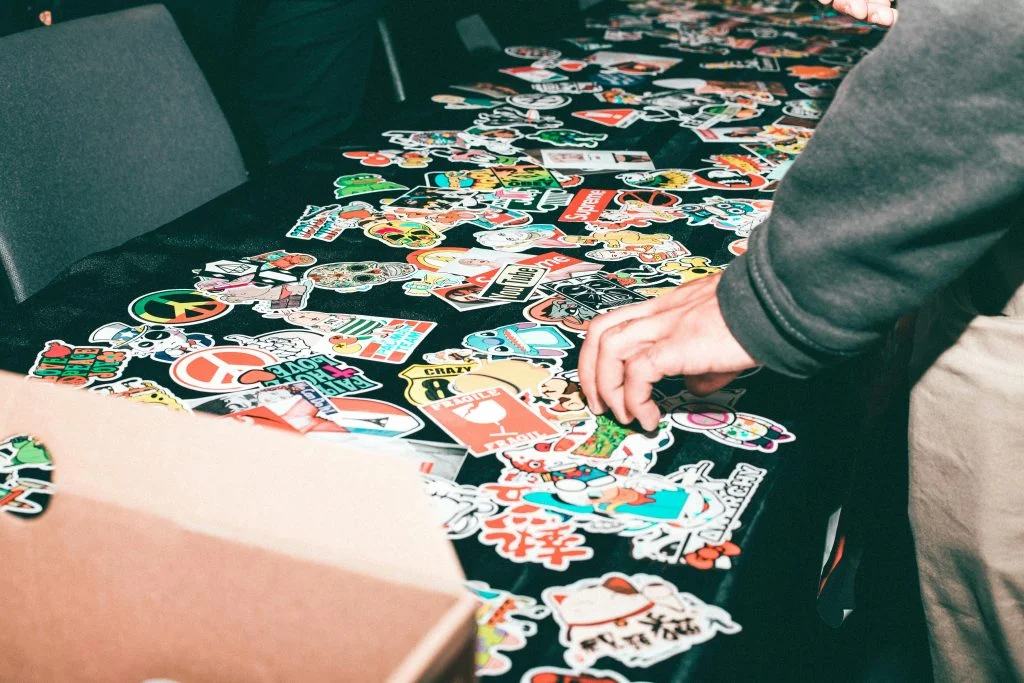 A person selecting from a large collection of colorful custom stickers displayed on a table, representing creative marketing and branding ideas for businesses.