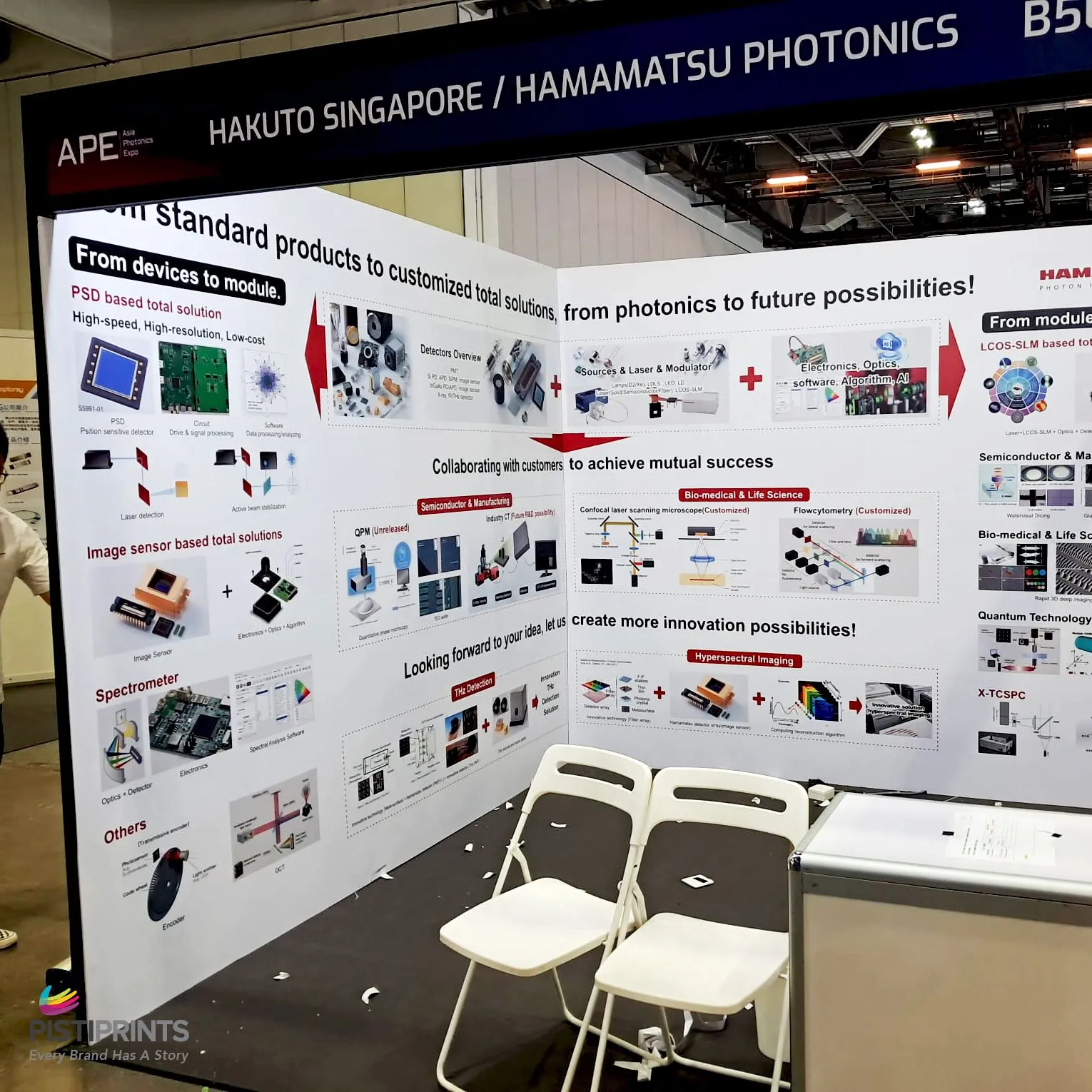 Exhibition booth of Hakuto Singapore and Hamamatsu Photonics at APE event, featuring printed backdrop panels showcasing photonics products, imaging solutions, and semiconductor technologies with chairs and display counter setup.