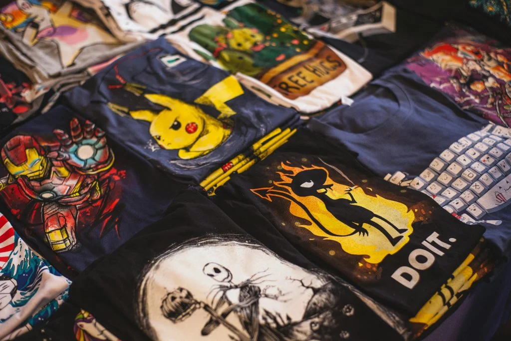 A collection of colorful branded T-shirts featuring popular cartoon and comic characters, neatly displayed on a table, representing promotional apparel for businesses.